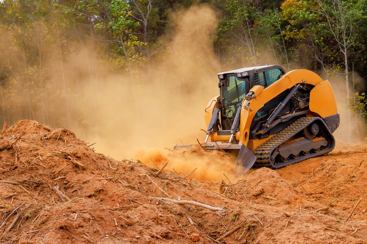 Skid steer clearing a plot of land in a forest, kicking up dust while moving soil